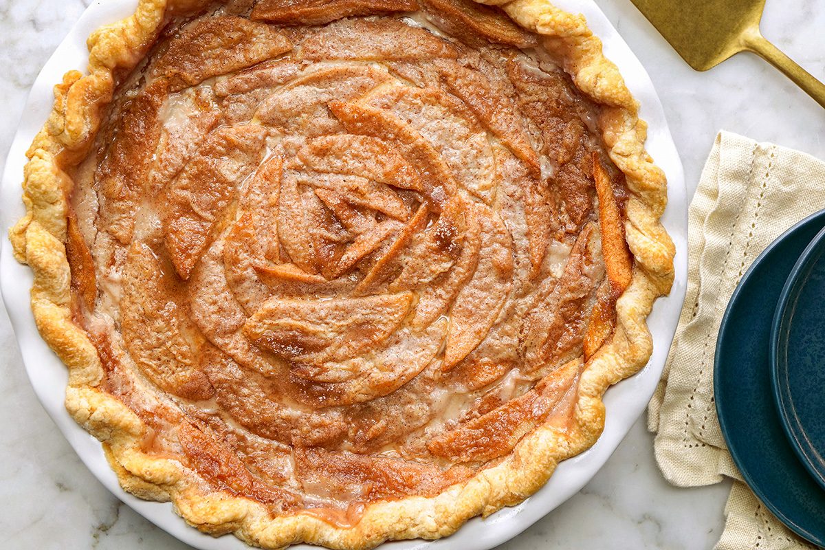 A baked peach pie with a golden, flaky crust and thinly sliced peach filling arranged in a spiral pattern sits in a white pie dish on a marble surface beside a gold server and dark blue plate.