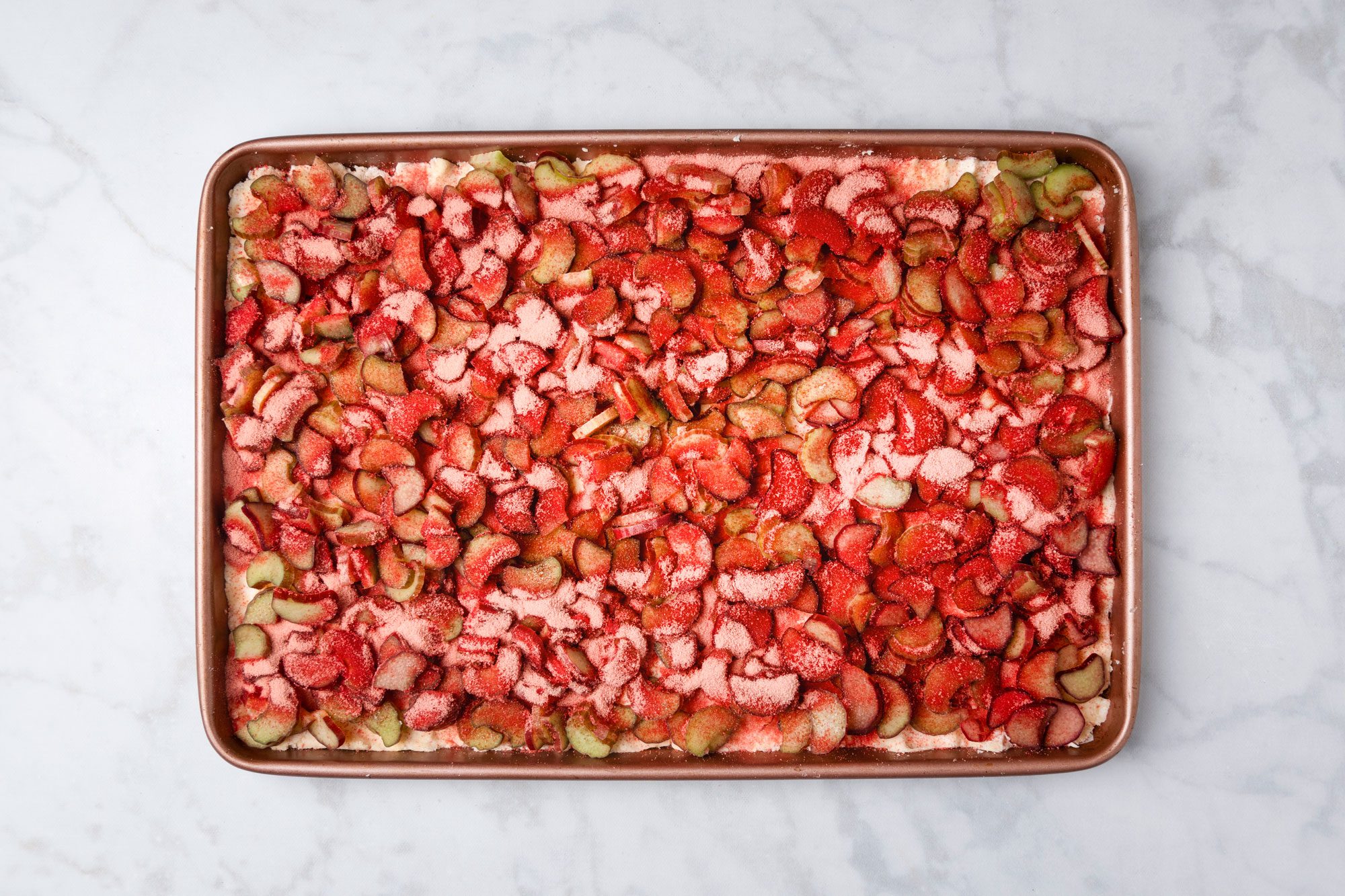 overhead shot of a baking sheet filled with an uncooked dessert topped with chopped strawberries and rhubarb, set on a light marble surface