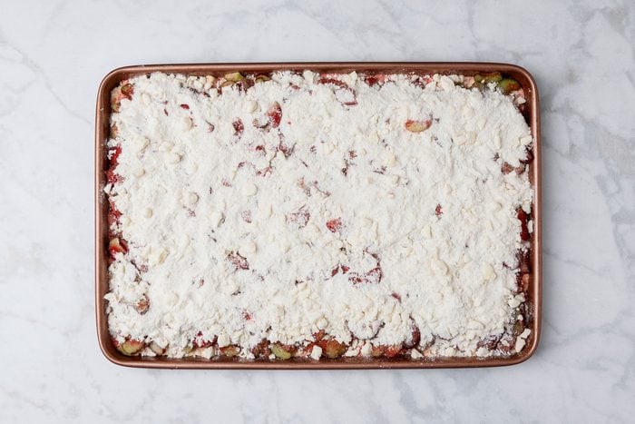 overhead shot of a rectangular baking sheet filled with an unbaked dessert, topped with a thick, crumbly layer of white flour mixture, Bits of fruit are partially visible underneath, The sheet is on a light marble surface