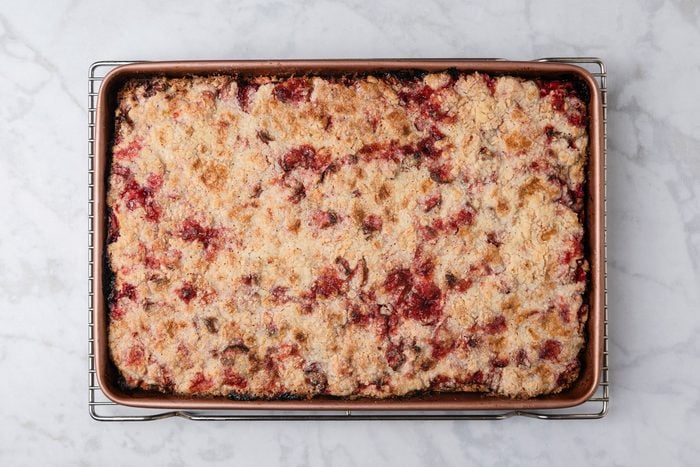 overhead shot of a rectangular baking pan filled with a golden brown, crumbly dessert topped with red fruit, possibly a berry crumble or crisp, sits on a wire rack on a white marble surface