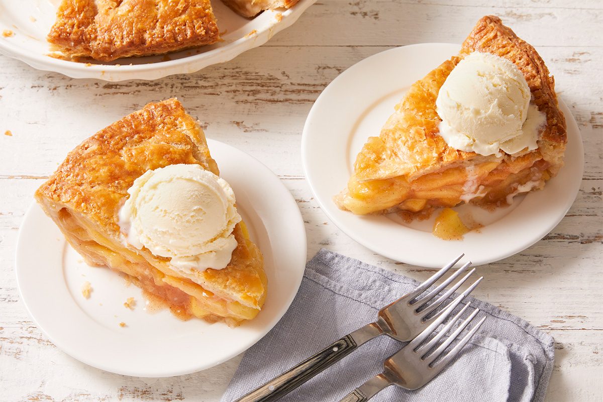 Two slices of apple pie topped with vanilla ice cream are served on white plates, accompanied by two forks on a folded blue napkin on a rustic white wooden table.