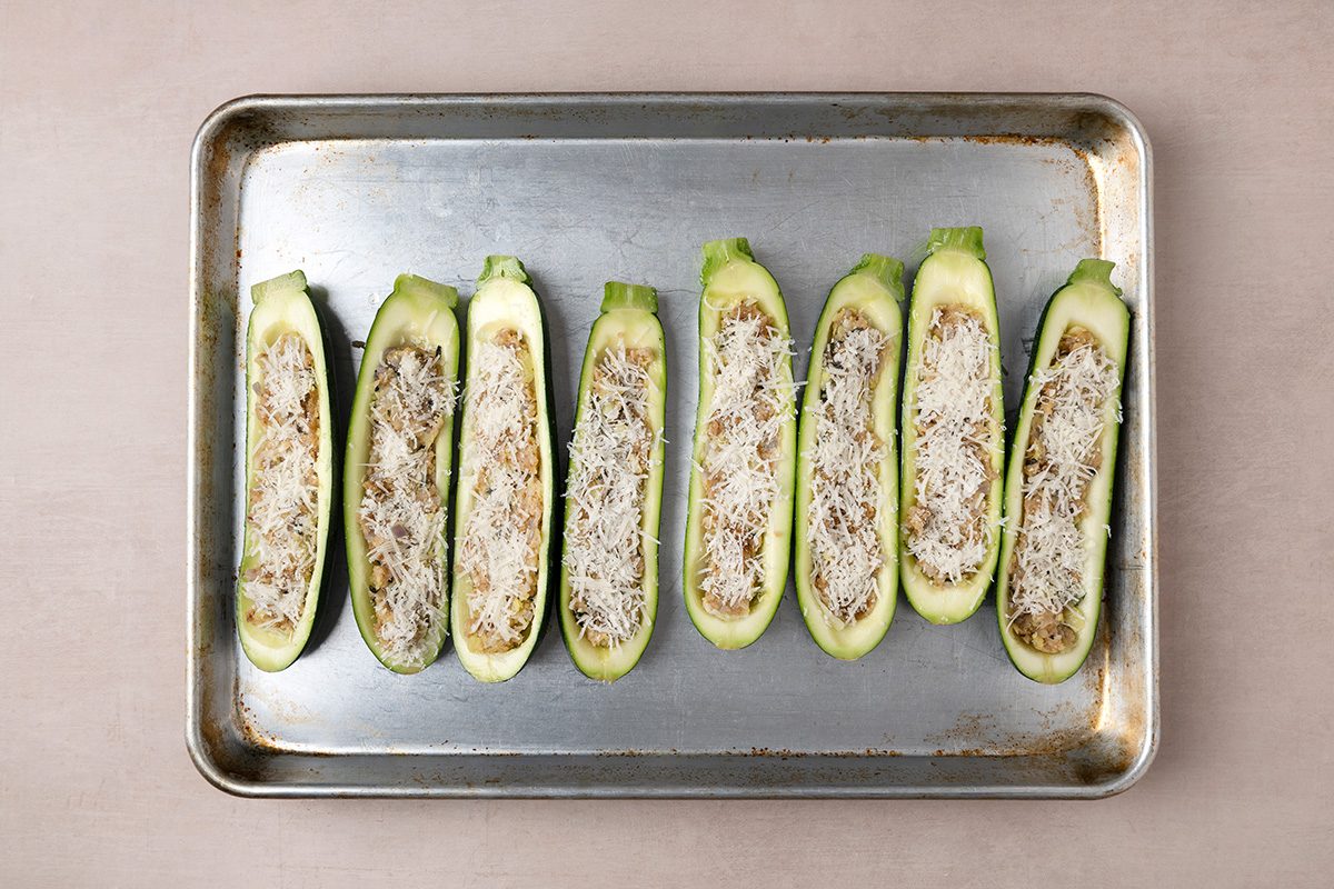 Eight zucchini halves, hollowed out and filled with a stuffing mixture, are arranged in a row on a metal baking sheet. The tops appear to be sprinkled with grated cheese.