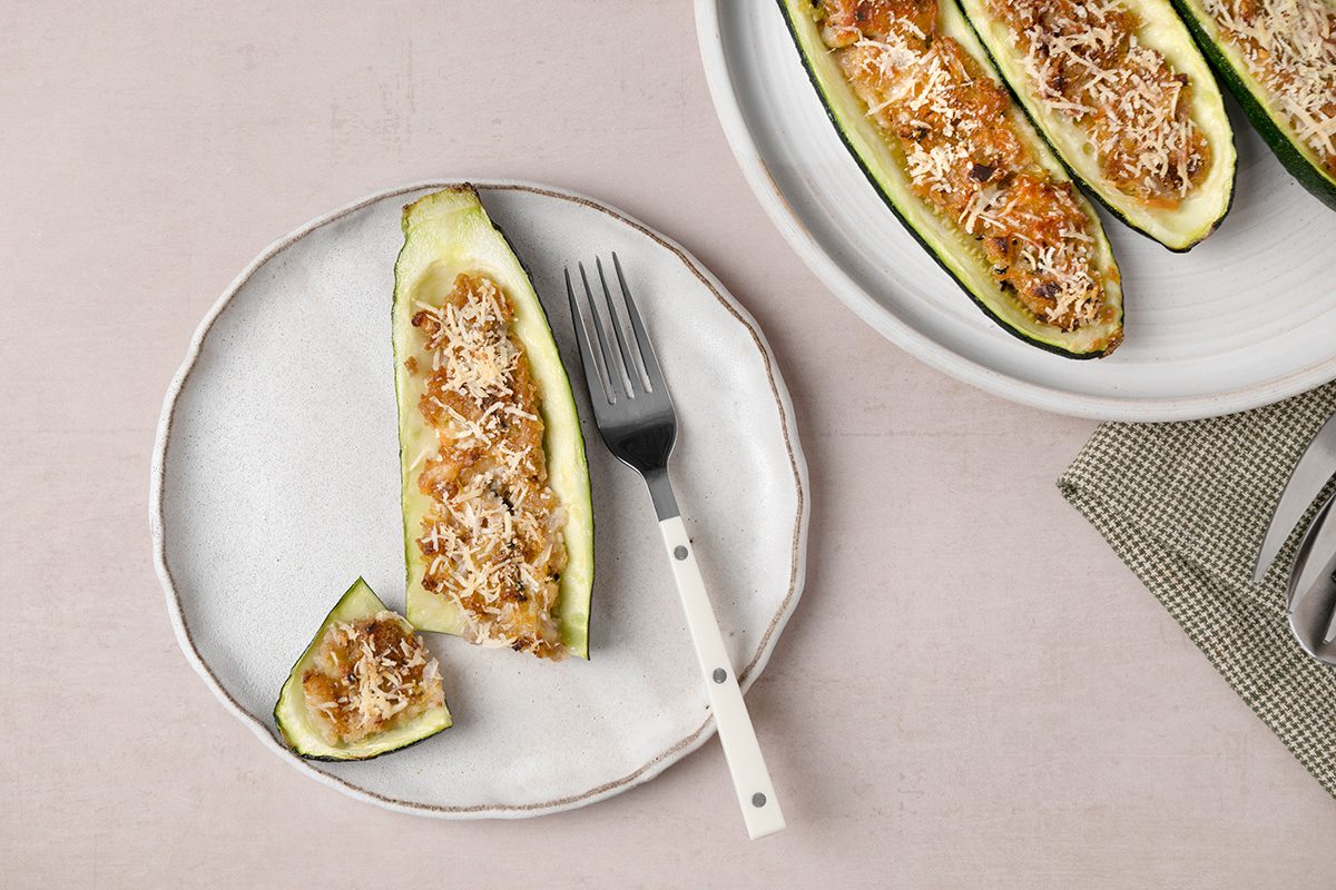 A stuffed zucchini boat topped with grated cheese sits on a white plate with a fork, while more stuffed zucchinis are visible on a larger plate nearby.