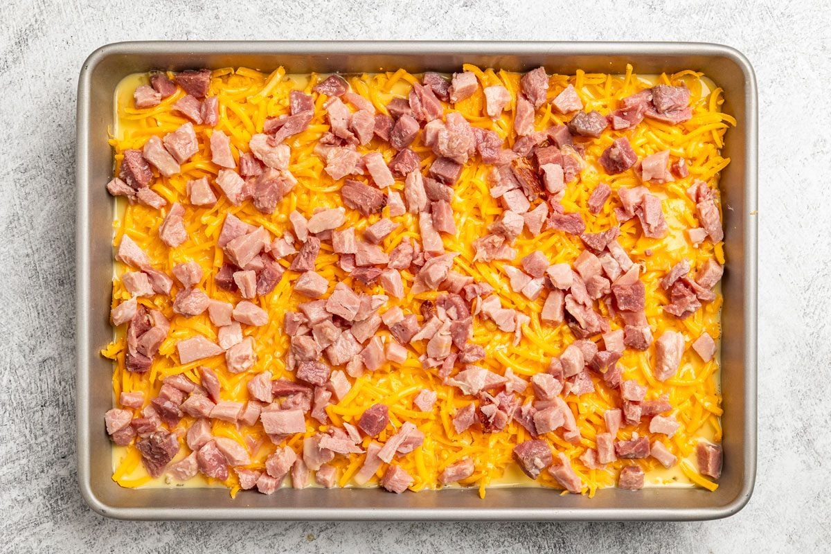 overhead shot of a metal baking tray filled with a layer of Hash Brown Patty Casserole and diced cooked ham on a light colored surface
