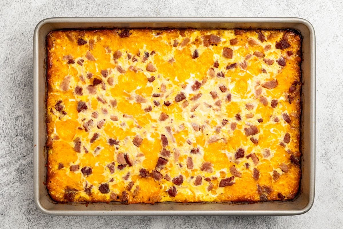 overhead shot of a rectangular casserole dish filled with a baked egg mixture, featuring chunks of cheese and pieces of ham throughout, sits on a light gray surface
