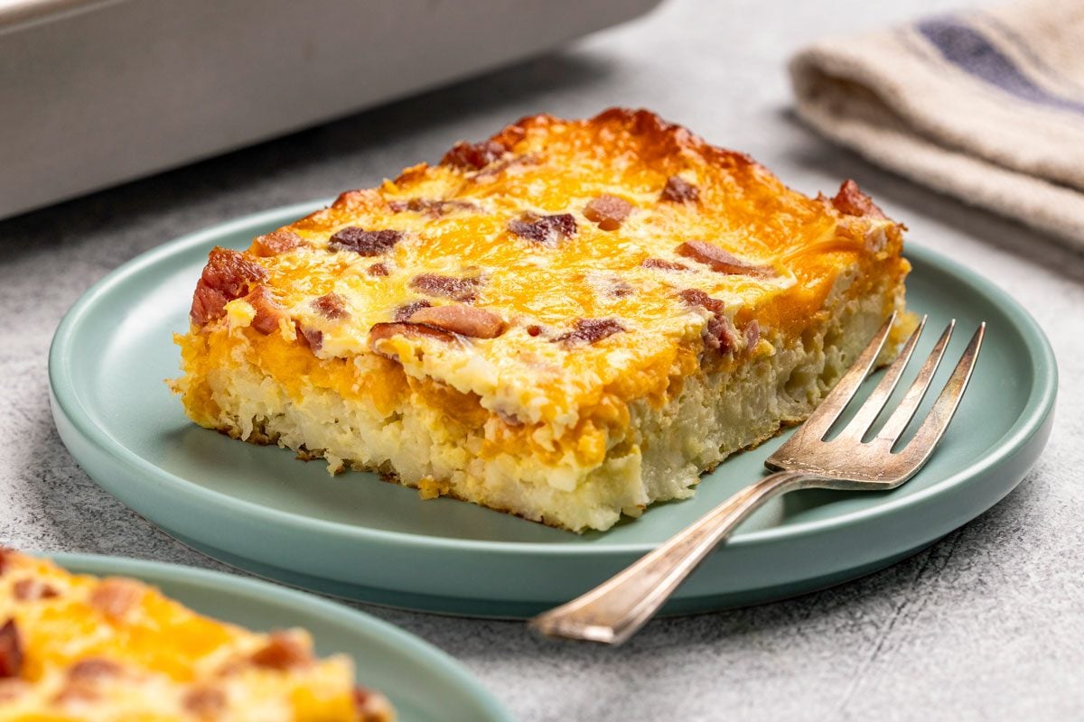 closeup shot of a slice of Hash Brown Patty casserole with eggs, cheese, and bacon sits on a light blue plate next to a fork, The casserole has a golden brown top and a layered texture