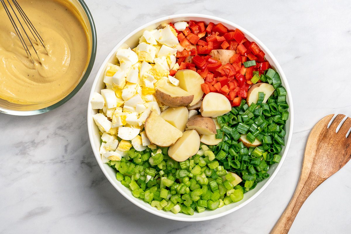 A bowl filled with chopped hard-boiled eggs, red bell pepper, green onions, celery, and potato pieces, next to a wooden utensil and a bowl of creamy dressing with a whisk on a marble surface.