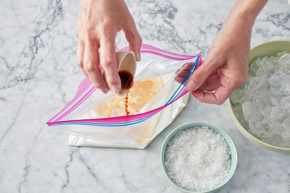 A person pours vanilla extract into a zip-top bag containing a creamy mixture. Nearby are bowls of ice and coarse salt on a marble countertop.