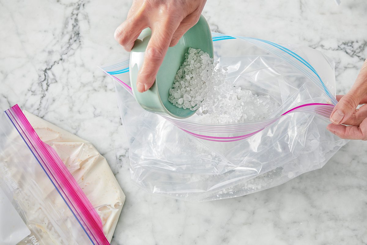 A person pours ice cubes from a green cup into a clear resealable plastic bag on a marble countertop. Another resealable bag with a light-colored substance is nearby.