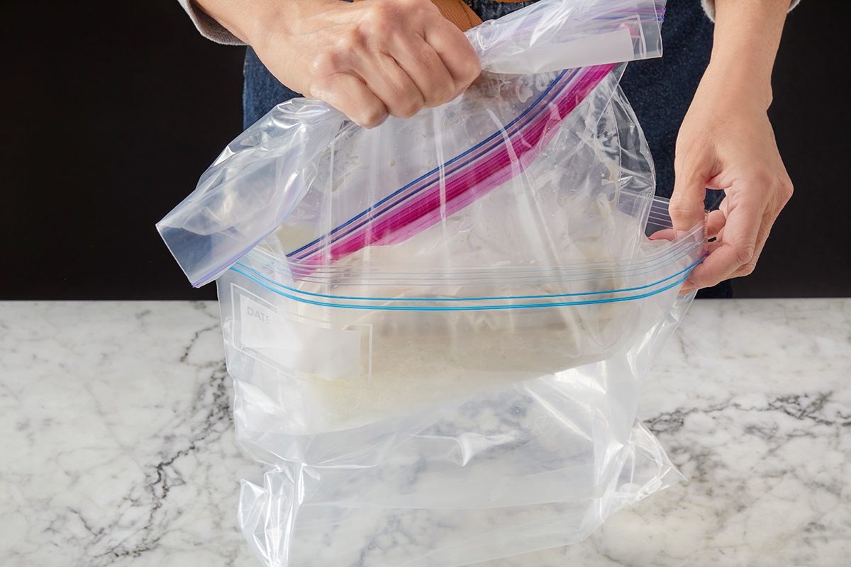 A person places a rectangular dish inside a large clear zip-top plastic bag on a marble countertop, preparing to seal the bag.