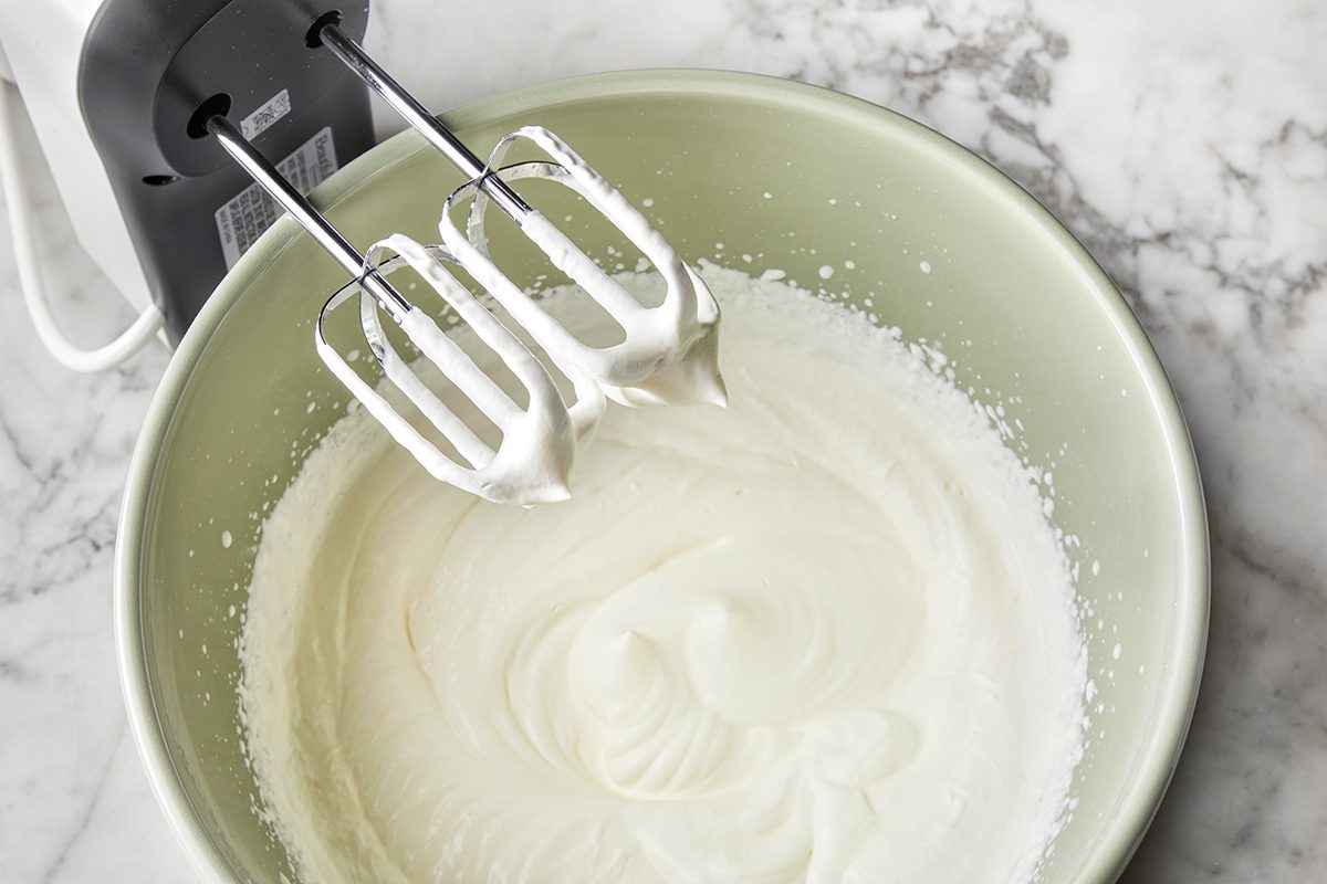 An electric hand mixer with beaters sits above a mixing bowl filled with freshly whipped cream on a marble countertop.