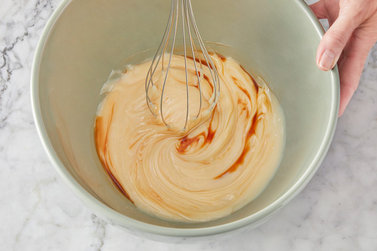 A hand holds a bowl while whisking a creamy mixture with swirls of vanilla extract on a marble countertop.