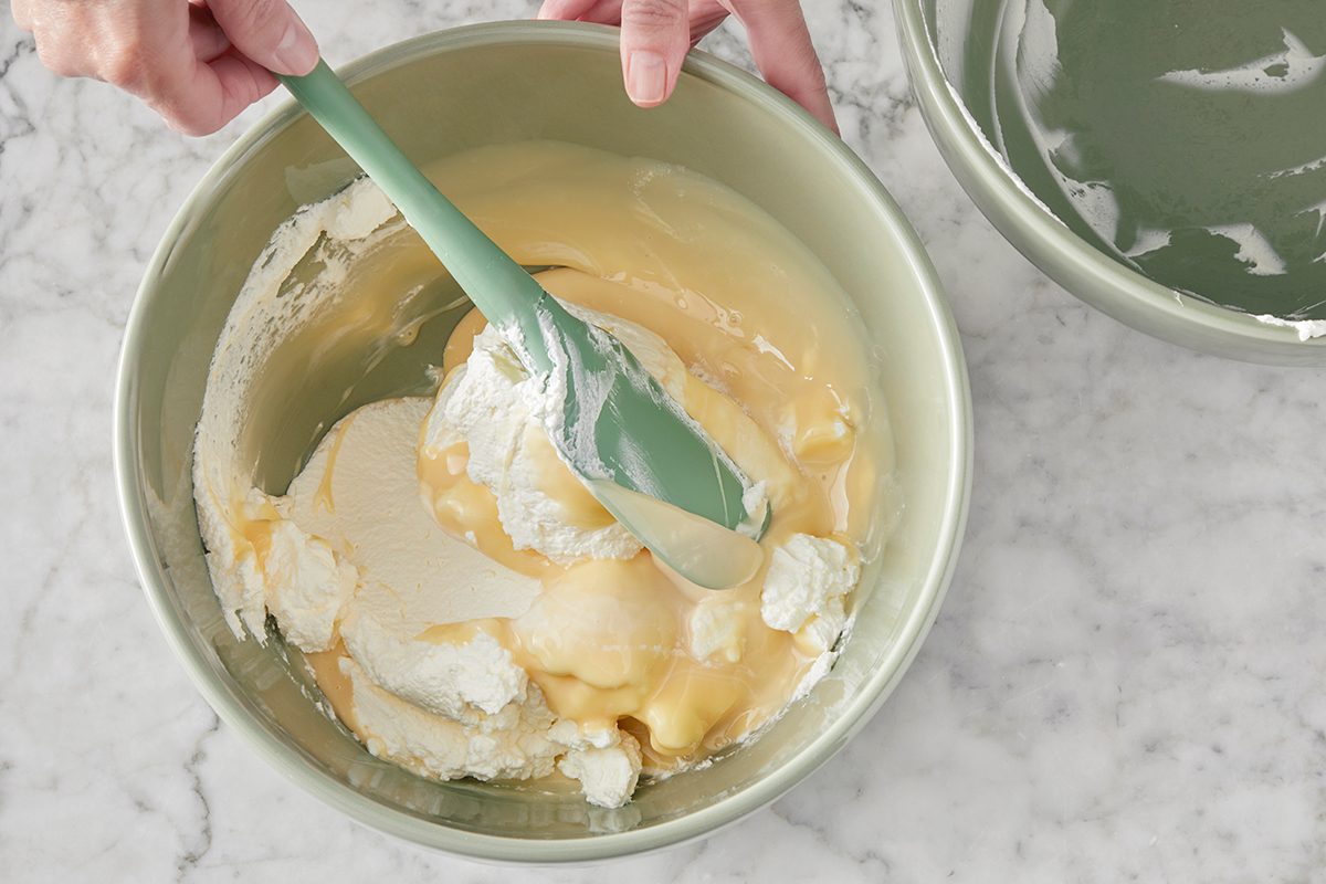 A person uses a green spatula to mix creamy yellow and white ingredients in a light green bowl on a marble countertop. Another empty bowl sits nearby.