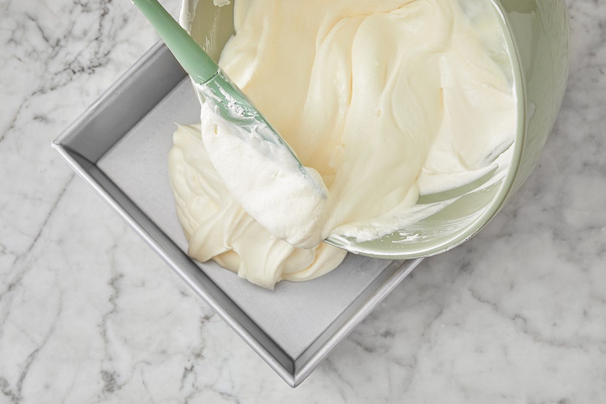 A mixing bowl pours thick, creamy batter into a square, parchment-lined baking pan on a marble countertop.