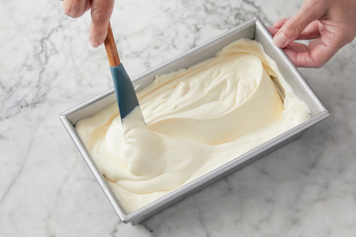 A person spreads creamy batter evenly in a rectangular metal baking pan using a blue silicone spatula on a marble countertop.
