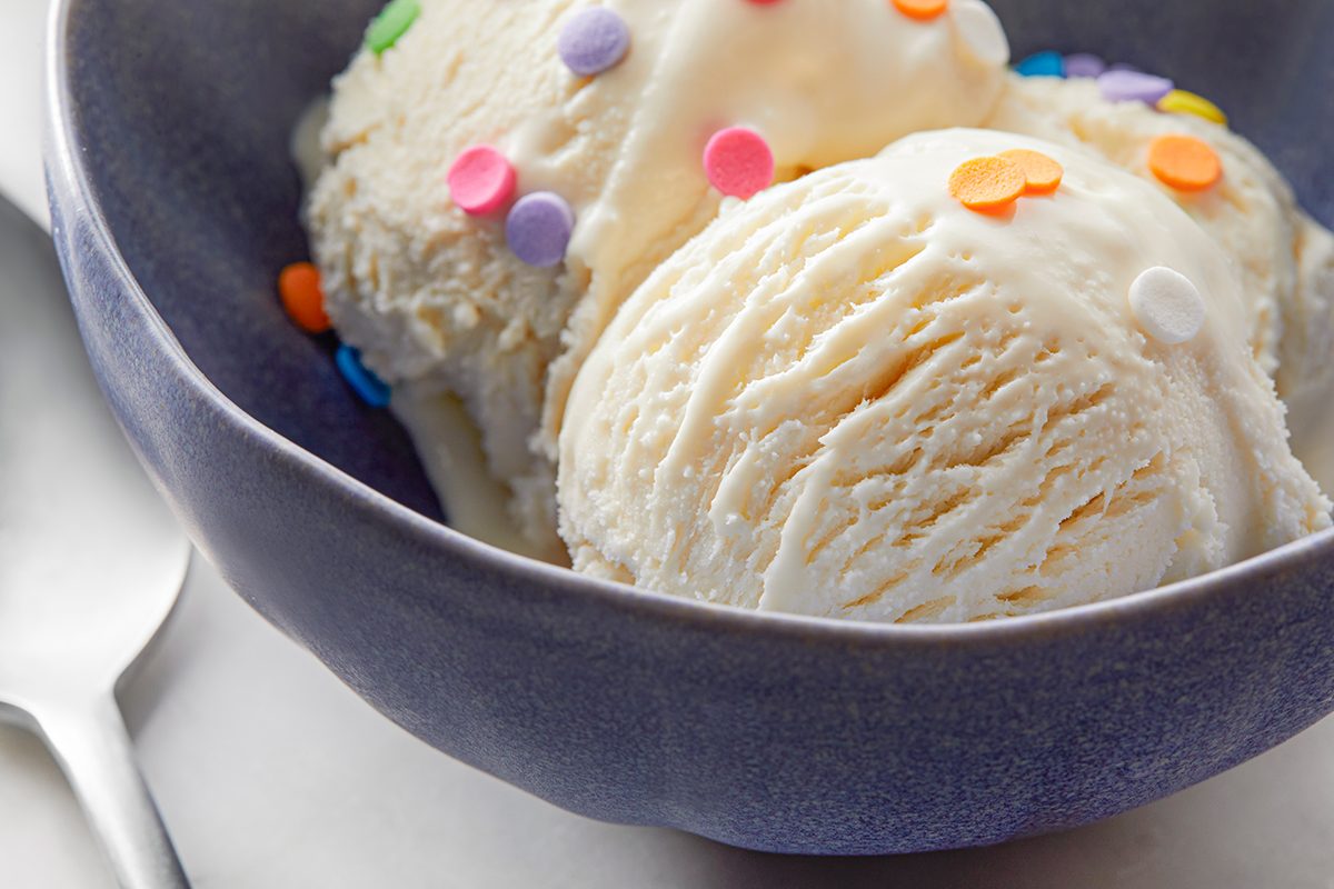 A close-up of two scoops of vanilla ice cream in a blue bowl, topped with colorful round sprinkles. A spoon is placed on the table beside the bowl.