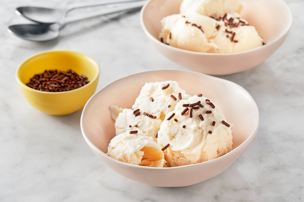 Two bowls of vanilla ice cream topped with chocolate sprinkles sit on a marble surface, with a small yellow bowl of extra sprinkles and two metal spoons in the background.