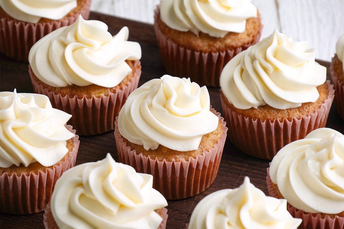 A group of vanilla cupcakes in brown paper liners topped with swirls of creamy white frosting, arranged closely together on a dark wooden surface.