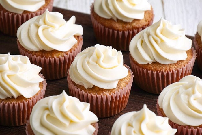 A group of vanilla cupcakes in brown paper liners topped with swirls of creamy white frosting, arranged closely together on a dark wooden surface.