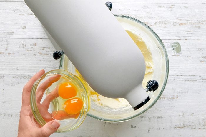 A hand holds a small glass bowl with two raw eggs above a stand mixer containing a light batter, ready to add the eggs. The scene is set on a white wooden surface.