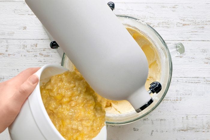 A person pours mashed bananas from a white bowl into a stand mixer with batter in a glass mixing bowl, preparing a recipe on a white wooden surface.