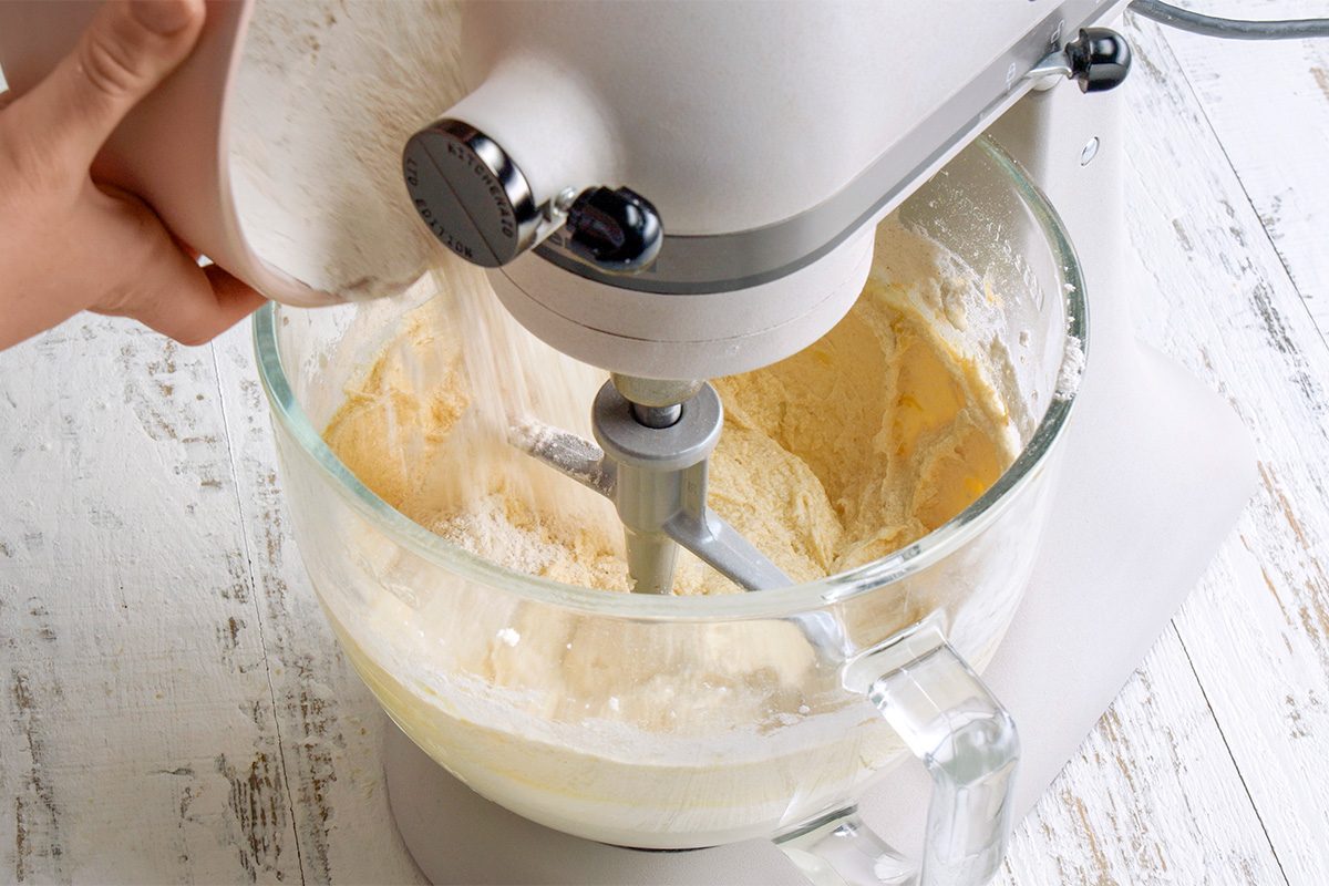 A person pours flour into a stand mixer bowl containing batter, with the mixer blending ingredients. The scene is set on a rustic white wooden surface.
