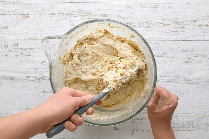 A person mixes dough with a spatula in a clear glass bowl on a white wooden surface.