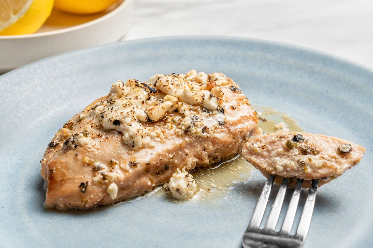 Close up shot of Lemon Feta Chicken served on a plate with a fork; slices of lemon in a bowl visible nearby; all set on a white marble;