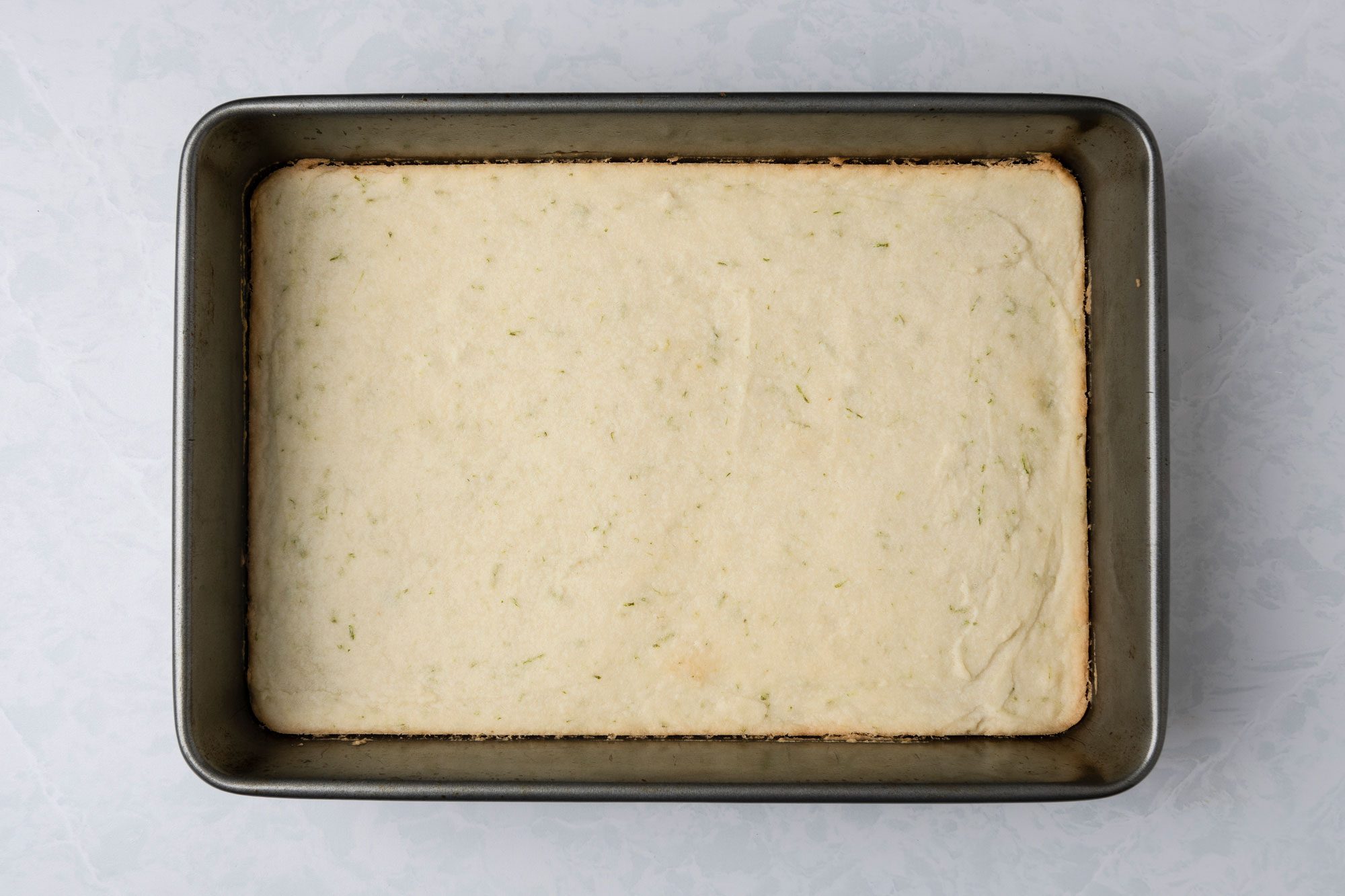 A top shot of a freshly baked sheet cake, slightly golden with a smooth top and tiny specks, rests in a rectangular pan on a light marbled background; signaling simplicity and freshness