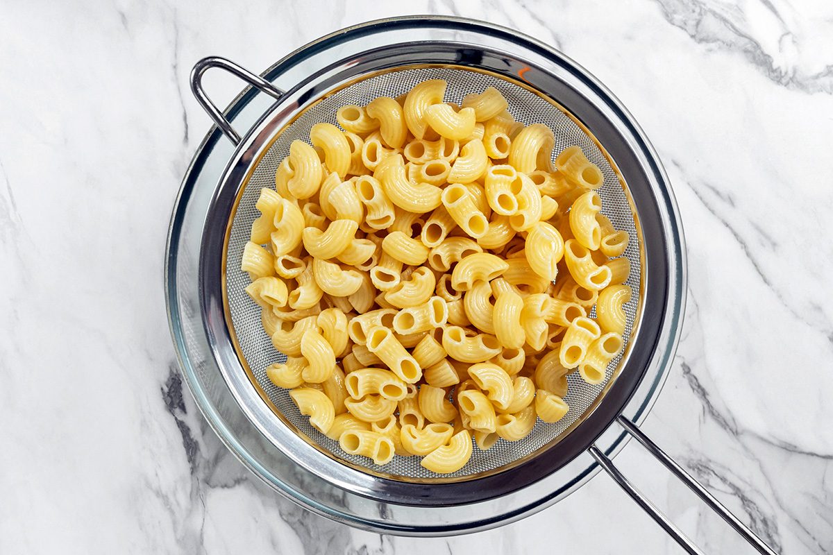 Cooked elbow macaroni pasta is draining in a metal colander placed over a bowl on a marble countertop.