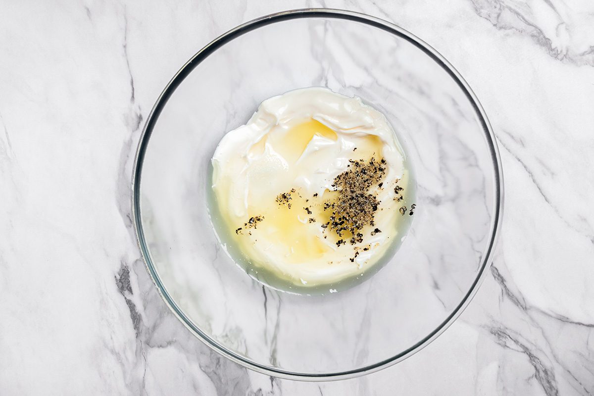 A clear glass bowl on a marble surface containing yogurt, olive oil, and dried herbs, ready to be mixed.