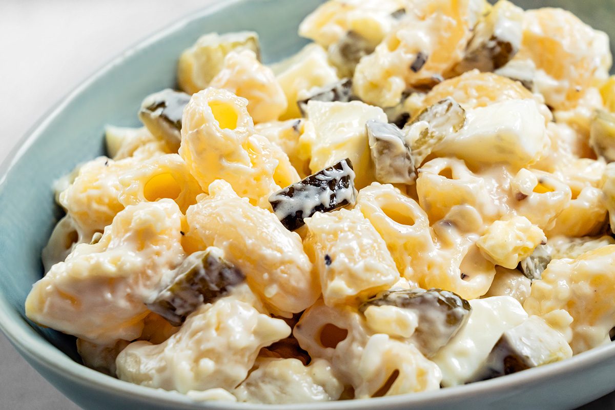 A close-up of a bowl filled with creamy macaroni salad, featuring pasta, diced pickles, chunks of cheese, and a mayonnaise-based dressing.