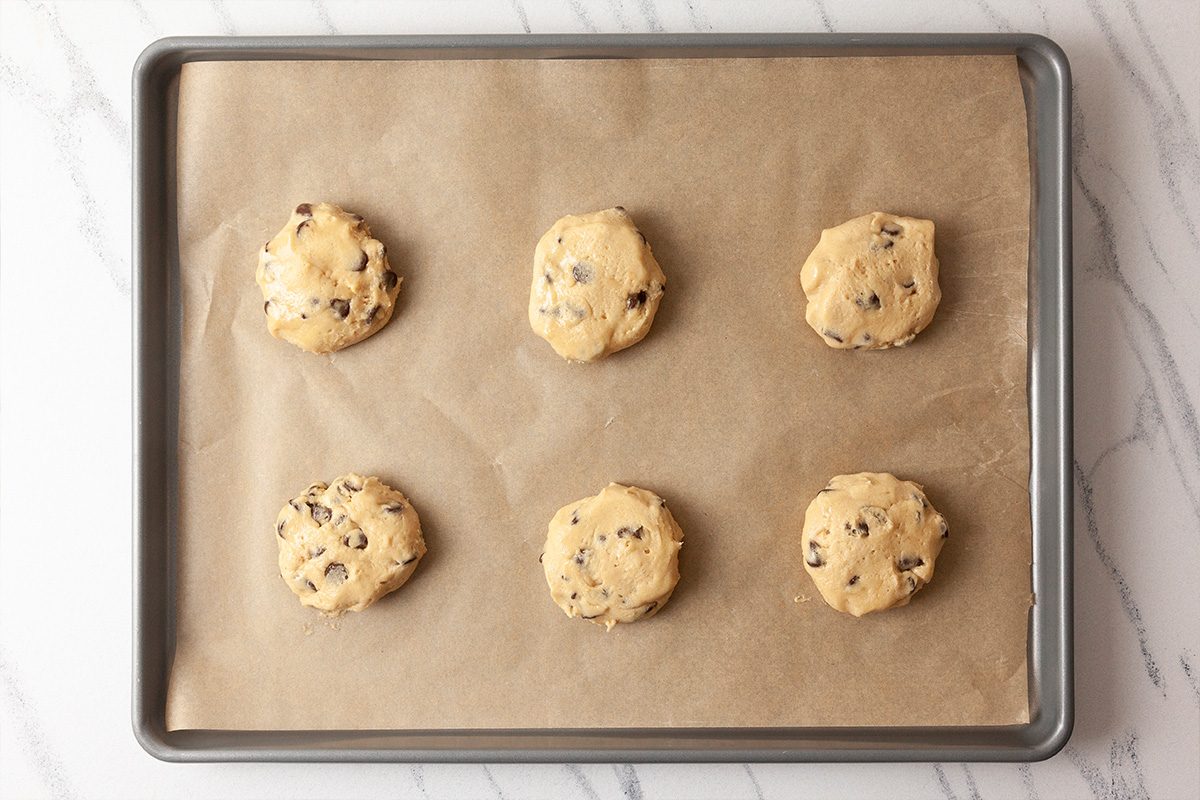 A baking sheet lined with parchment paper holds six evenly spaced, unbaked chocolate chip cookie dough balls on a marble surface.