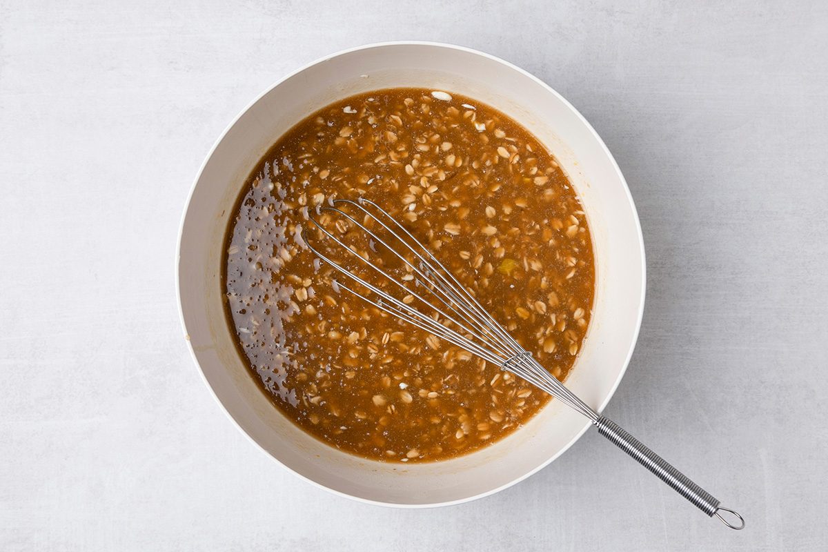 A metal whisk rests in a white bowl filled with a chunky brown sauce or mixture, set on a light gray surface.