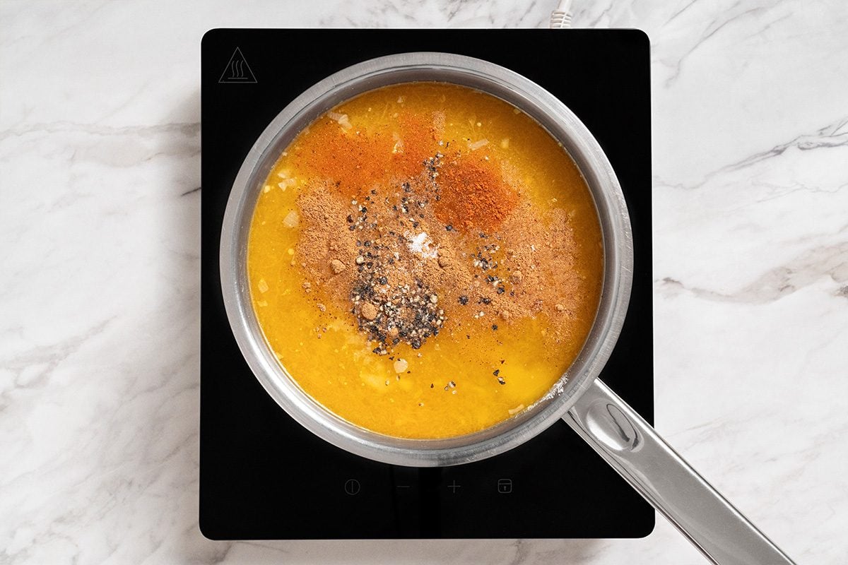 A saucepan filled with yellow soup or sauce topped with various spices sits on a black electric stovetop on a white marble surface.