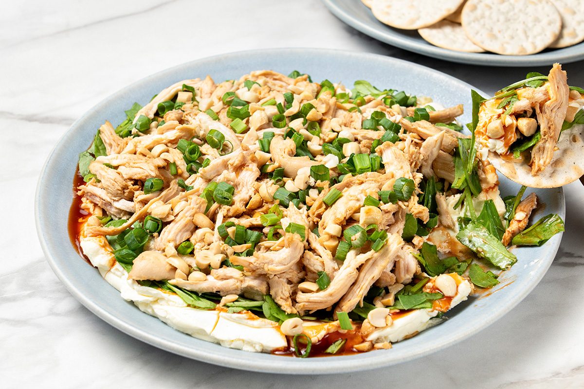 A plate of shredded chicken salad mixed with green onions and sauce, served on a bed of leafy greens with a side of round crackers on a marble surface.