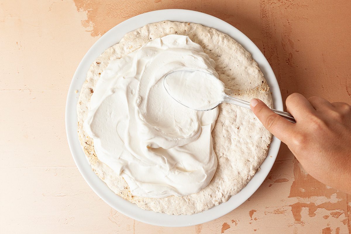 A hand uses a spoon to spread a thick layer of white cream or frosting over a round cake layer on a white plate, set on a light-colored surface.