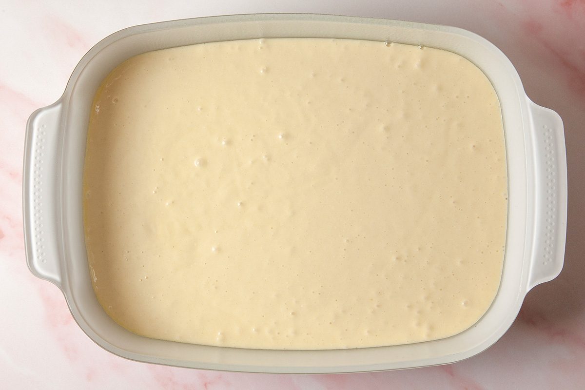 overhead shot of a white rectangular baking dish filled with smooth, pale yellow cake batter sits on a light pink marble surface, ready to be baked