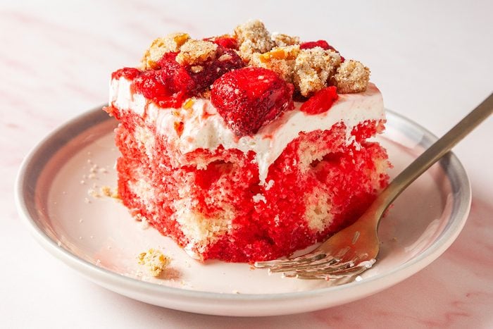 3/4th closeup shot of a slice of strawberry shortcake with whipped cream, strawberry topping, and crumbly streusel, served on a white plate with a fork