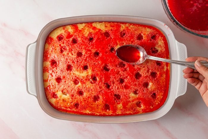 overhead shot of a hand holds a spoon, pouring red liquid into holes in a baked yellow cake in a white rectangular dish; The cake has evenly spaced holes and sits on a pink marble countertop; A bowl of red liquid is nearby