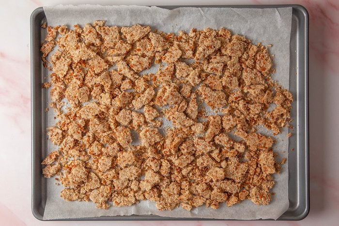overhead shot of a baking sheet lined with parchment paper is covered with an even layer of crumbled, toasted brown bread pieces on a light pink marble surface
