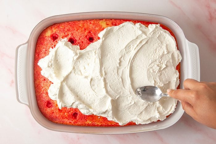 overhead shot of a hand spreads white whipped topping over a rectangular strawberry poke cake in a white baking dish, using a spoon; The cake has red spots where strawberry syrup was poured into hole