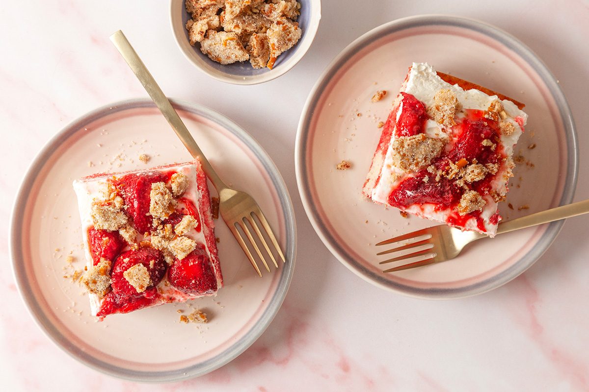 overhead shot of Two plates each hold a square slice of strawberry shortcake topped with whipped cream and crumbled cookie, with gold forks beside them; A small bowl of extra crumb topping is above the plates on a pink marble surface
