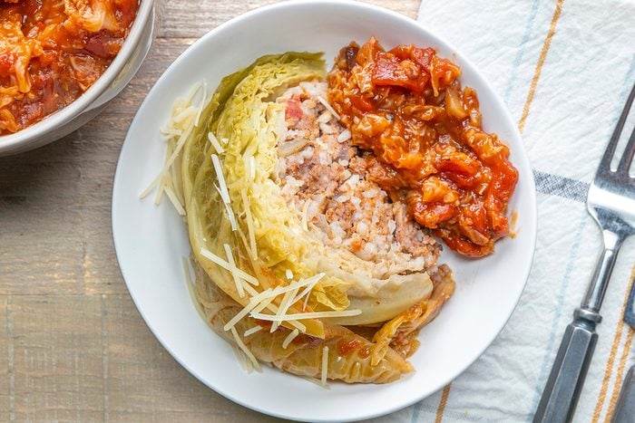A white plate with a wedge of stuffed cabbage filled with rice and meat, topped with tomato sauce and sprinkled with grated cheese, next to a fork and knife on a white napkin.