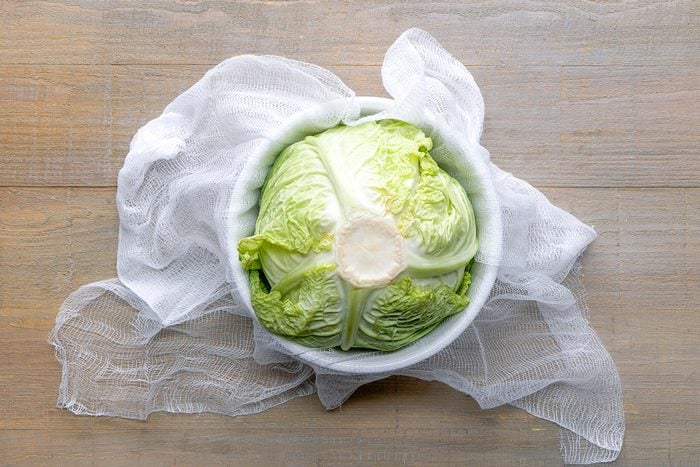 A whole head of napa cabbage sits in a white bowl, placed on a light wooden surface with a white cloth underneath.