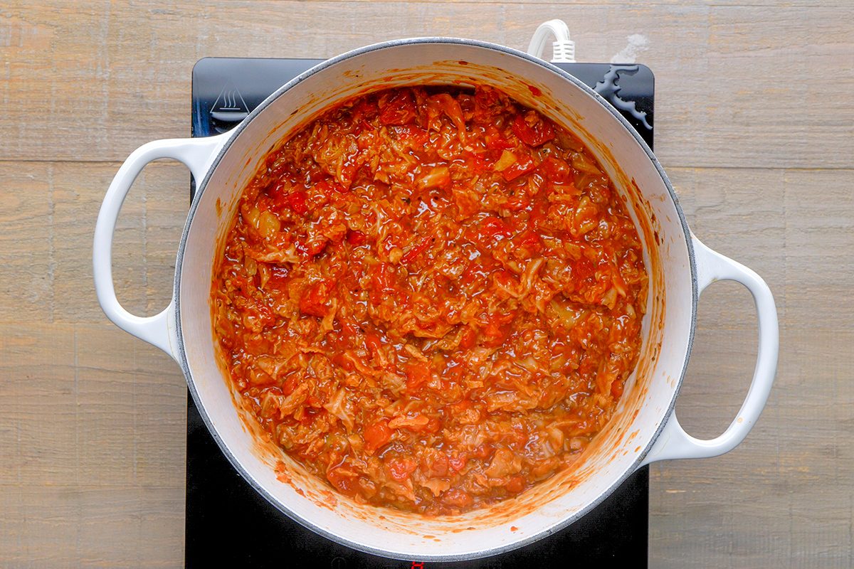 A white pot filled with thick, chunky red tomato sauce simmers on a black stovetop, with visible bits of vegetables and herbs throughout.