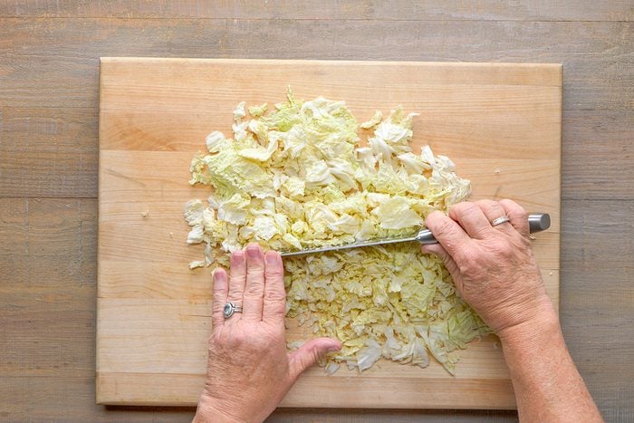 A person’s hands chopping leafy cabbage on a wooden cutting board with a knife on a light wood table.