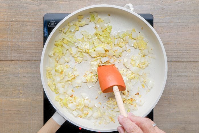 A hand stirs chopped onions cooking in a white frying pan with an orange spatula on a stovetop.