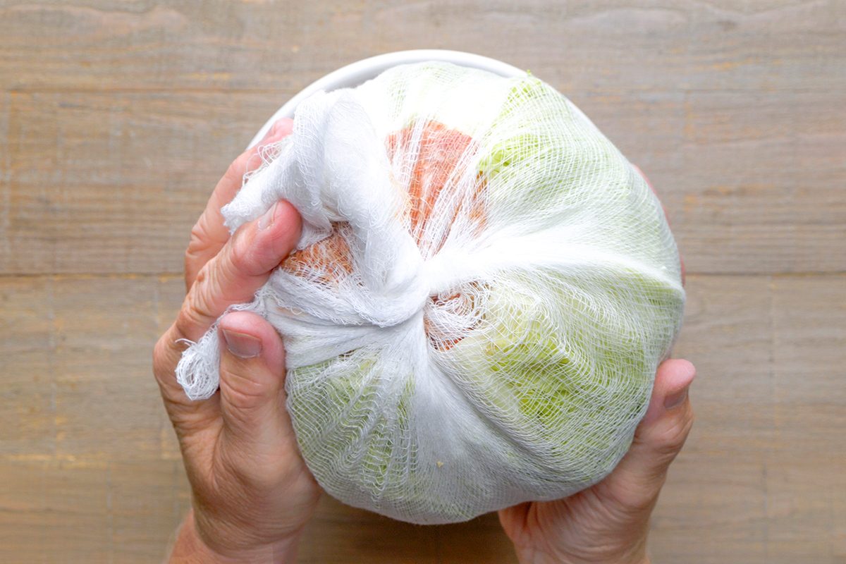 Two hands holding a bundle of food items wrapped tightly in white cheesecloth against a light wooden background.