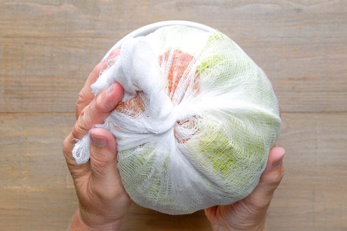 Two hands holding a bundle of food items wrapped tightly in white cheesecloth against a light wooden background.